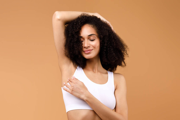 Hair removal concept. Happy latin lady demonstrating her armpit with smooth skin and smiling, posing in white underwear over brown background, copy space - Foto, imagen