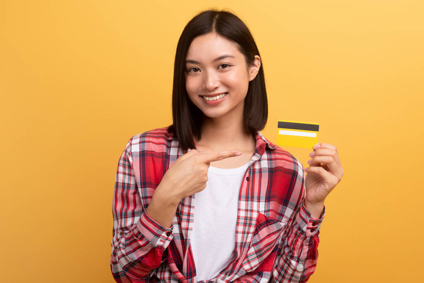 Cheerful japanese lady shopaholic pointing finger at credit card, recommending sale isolated on yellow background. Finance and money for shopping, ad and offer - Photo, Image