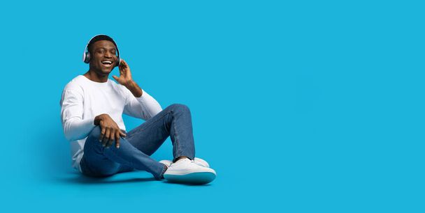 Fajna playlista. Szczęśliwy przystojny młody Afroamerykanin w luźnym siedzeniu na podłodze, słuchając muzyki na niebieskim tle studio, za pomocą bezprzewodowych słuchawek i uśmiechnięty, panorama z miejsca kopiowania - Zdjęcie, obraz