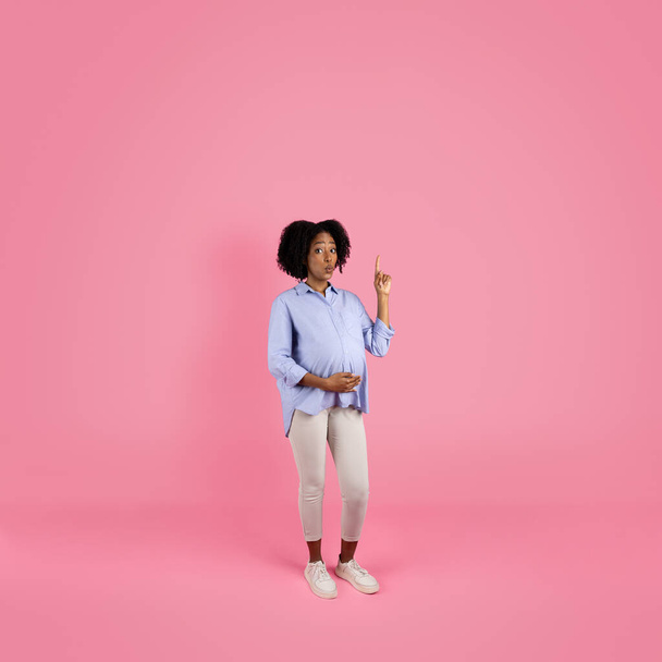 Smiling shocked inspired young black pregnant woman in casual touching big belly with hand, pointing finger at empty space, isolated on pink studio background. Got idea, ad and offer, waiting baby - Fotó, kép