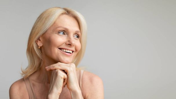 Attractive middle aged woman with hydrating glowing skin looking aside at free space and smiling, posing on grey studio background, panorama. Modern cosmetology concept - Foto, imagen