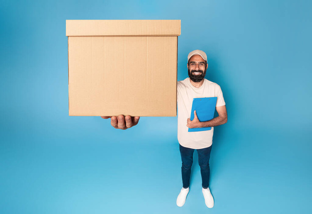 Entrega indiana homem segurando caixa de papelão pacote que se estende para a câmera, de pé sobre fundo azul e sorrindo, vista acima. Transporte e prestação de serviços - Foto, Imagem