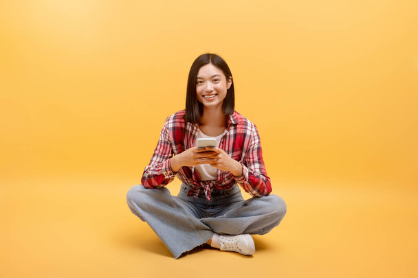 Onnellinen aasialainen nainen, joka käyttää kännykkää tekstiviestillä tai selailee Internetiä, istuu lattialla keltaisen taustan päällä hymyillen kameralle, vapaa tila. Matkapuhelimen mobiilisovellus - Valokuva, kuva