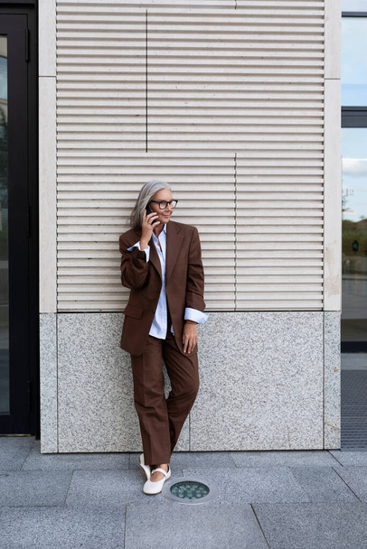 egy sikeres ősz hajú üzleti nagymama öltözött elegáns barna öltöny megállt a falon az üzleti központ használni az okostelefon. - Fotó, kép
