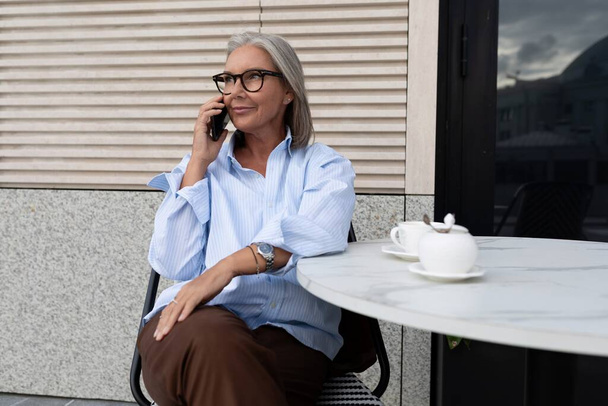 successful free gray-haired business grandmother dressed in a stylish shirt sits on the terrace of a cafe and speaks on the phone. - Photo, Image