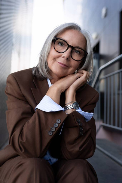 well-groomed healthy slender gray-haired mature business lady dressed in a stylish brown jacket sits against the backdrop of a business building. - Foto, Bild