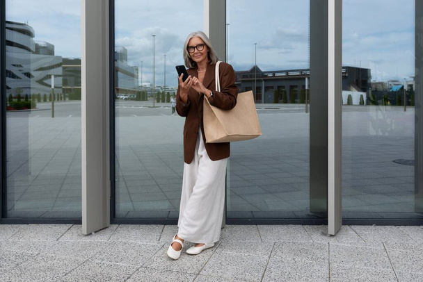 uma senhora de negócios madura de cabelos grisalhos vestida com uma jaqueta elegante e calças está de pé na entrada do centro de negócios. - Foto, Imagem