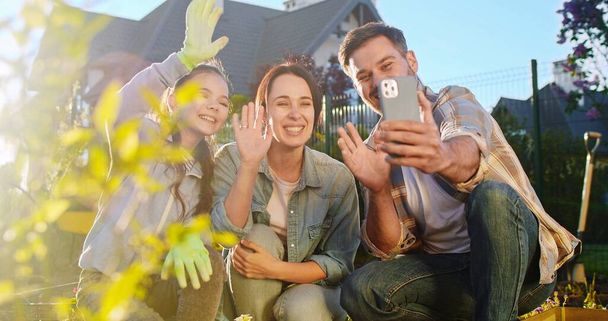 Cheerful Caucasian family with flowers in pots smiling to camera and videochatting on smartphone. Working in orchard. Father, moher and daughter planting garden and having videochat via mobile phone. - 写真・画像