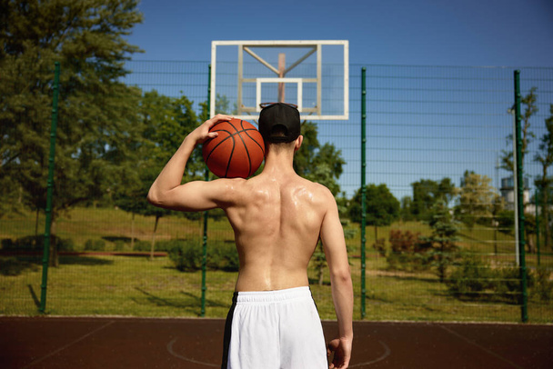 Pohled zezadu na svalnaté tělo basketbalového hráče bez košile, který drží míč na rameni. Sportovní trénink venku - Fotografie, Obrázek