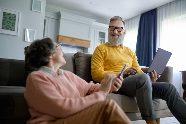 Χαμογελώντας συνταξιούχος ζευγάρι χρησιμοποιώντας ψηφιακό gadget ενώ στηρίζεται στο σαλόνι στο σπίτι. Ηλικιωμένη σύζυγος συνομιλεί με φίλους στο smartphone και ανώτερος σύζυγος μηνυμάτων μέσα κοινωνικής δικτύωσης - Φωτογραφία, εικόνα