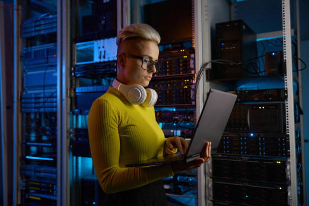 Vista lateral da mulher engenheira trabalhando no laptop fazendo operação de backups em equipamentos de rede na sala de servidores escuros - Foto, Imagem