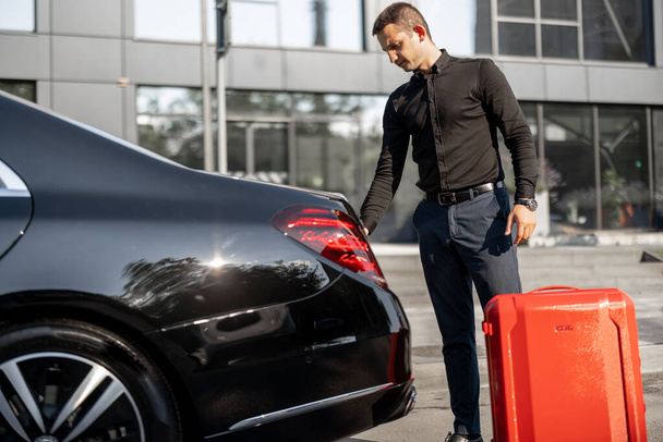 Водитель автомобиля бизнес-класса упаковывает красный чемодан в багажник на улице возле офисного здания. Концепция деловых поездок, личного водителя или роскошного такси - Фото, изображение