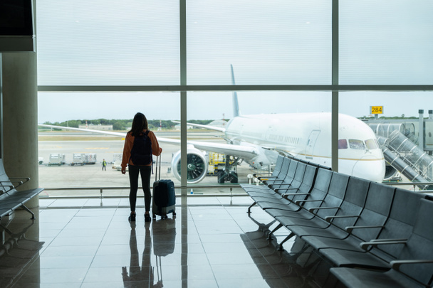 Mujer con la maleta en la mano, mira por la ventana del aeropuerto mientras espera su vuelo. - Foto, Imagen