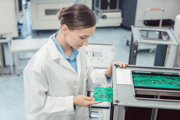 vrouw elektrotechnicus het dragen van een witte labjas werken bij assemblagelijn voor elektronische goederen het uitvoeren van optische controle op printplaten - Foto, afbeelding