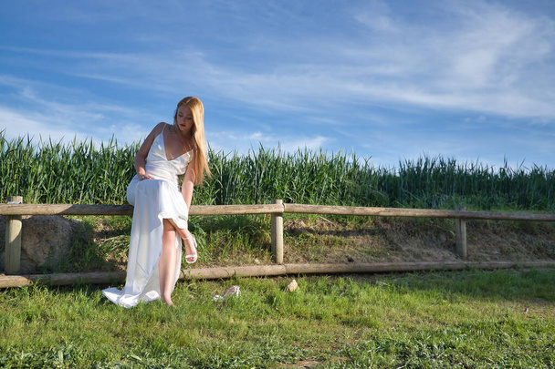 Молодая женщина, красивая и блондинка в белом платье, сидит на деревянных перилах, надевая туфли, с лугом на заднем плане. красота, мода, невесты, обувь, ноги, аксессуары. - Фото, изображение