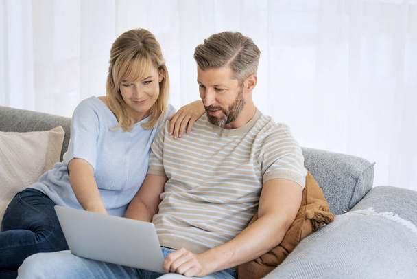 Middle-aged couple sitting at home on the sofa and using a laptop. Blond haired woman and grey haired man surfing the internet or having video call.  - Foto, immagini