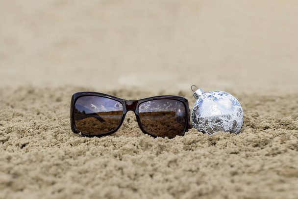 Kumsalda Noel. Güneş gözlüğü ve kumda bir Noel topu. Sıcak ülke konseptinde yeni yıl ve Noel tatili. - Fotoğraf, Görsel