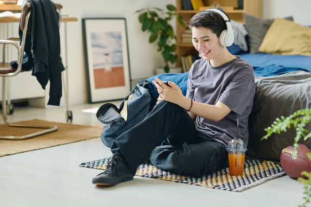 Χαριτωμένο χαμογελαστό μελαχρινή κοπέλα σε λευκά ακουστικά επικοινωνώντας σε συνομιλία βίντεο ή παίζοντας νέο παιχνίδι στον ελεύθερο χρόνο, ενώ κάθεται στο πάτωμα από το κρεβάτι - Φωτογραφία, εικόνα