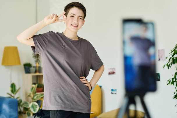 Menina feliz em camiseta cinza mostrando gesto de paz e olhando para a câmera do smartphone enquanto fotografa ao vivo para o público online - Foto, Imagem