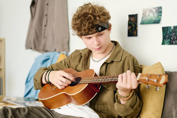 Teenage chlap s kudrnaté vlasy hrát akustickou kytaru při relaxaci na posteli nebo na gauči před kamerou a těší víkend doma - Fotografie, Obrázek
