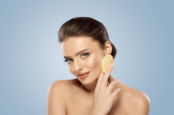 Attractive woman with makeup holding a cellulose sponge in her hand over a gray background with copy space. Face cleansing, makeup removing, skincare routine. Concept of wellness. Beauty blog - Zdjęcie, obraz