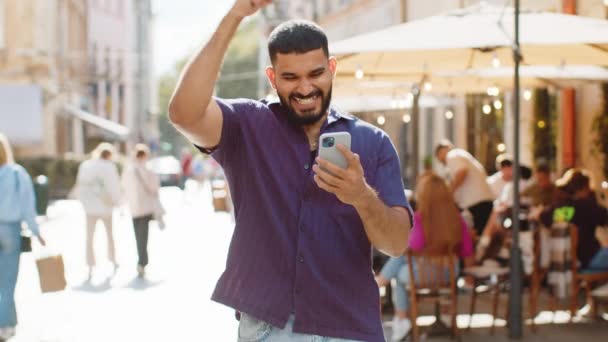 El joven indio utiliza el teléfono inteligente móvil celebrando ganar buenas noticias de mensaje, la victoria del bote de lotería, sorteo en línea al aire libre. Feliz adulto chico turista caminando en la calle urbana de la ciudad. Estilo de vida de la ciudad - Metraje, vídeo