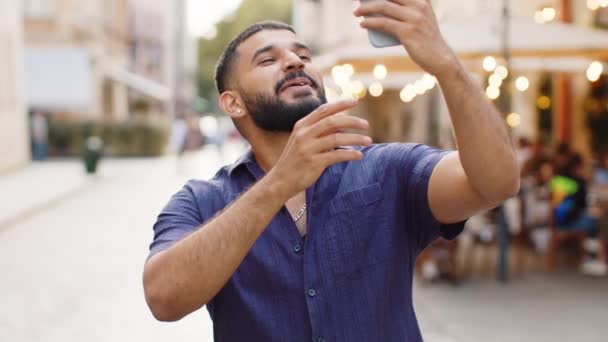 Brodaty blogger indiański człowiek robi selfie na smartfonie, komunikuje wideo rozmowy online z subskrybentami lub przyjaciółmi rodziny, nagrywa historie dla mediów społecznościowych vlog na świeżym powietrzu. Facet spacerujący ulicą miasta - Materiał filmowy, wideo