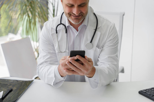 Улыбающийся доктор в форме сидит на рабочем месте в офисе во время перерыва и держит телефон  - Фото, изображение