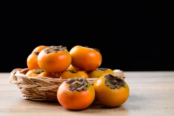 Fresh persimmon fruit in basket on wooden table with black background, Autumn seasonal fruit - Photo, Image