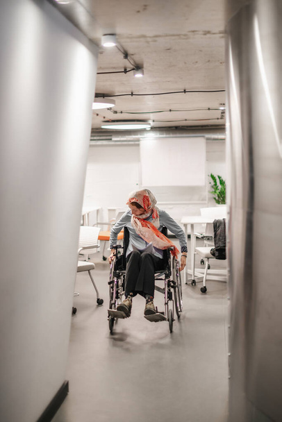 Donna disabile in hijab rotolando la sedia a rotelle attraverso l'ufficio al lavoro - Foto, immagini