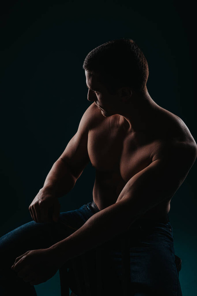 Jeune bodybuilder torse nu posant en studio, assis sur une chaise et regardant de côté. Silhouette photo - Photo, image