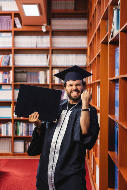 Graduado joven y emocionado con una gorra y un vestido mientras sostiene su título de licenciatura - Foto, imagen