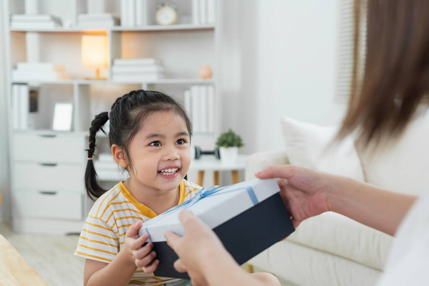 Grateful asian girl holding blue present box giving mothers day gift to mom. Happy mom and cute daughter kid child celebrating birthday, hugging on couch sofa at home. Happy family concept. - Photo, Image