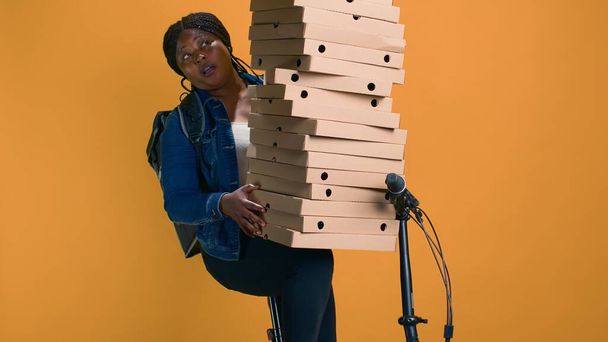 Junge schwarze Frau mit einem Stapel Pizzakartons für die Zustellung zu einer Feier in der Nachbarschaft. Afrikanischer Lieferant balanciert vorsichtig riesige Bestellung für Essenslieferung aus. - Foto, Bild