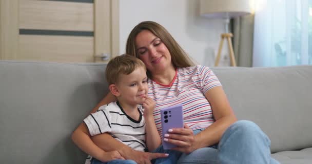 Woman and her little son sitting on cozy sofa with smartphone. Family spend leisure together at home using smartphone, play games, watch video, take selfies. High quality 4k footage - Filmati, video