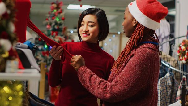 Cliente asiático interessado em blusa vermelha para usar durante o inverno festa temática festiva, perguntando empregado sobre materiais e preço. Cliente que precisa de traje de xmas para celebrações de Natal - Foto, Imagem