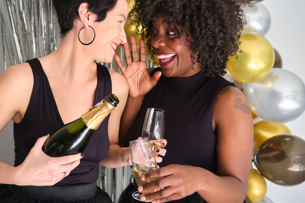 Δύο γυναίκες που σερβίρουν ποτήρια σαμπάνιας από το μπουκάλι και γιορτάζουν την Πρωτοχρονιά σε ένα πρωτοχρονιάτικο πάρτι. - Φωτογραφία, εικόνα