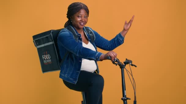 Γυναίκα Αφροαμερικάνα ποδηλάτισσα σε κίτρινο φόντο που κουβαλάει τσάντα παράδοσης τροφίμων στην πλάτη της. Μαύρη γυναίκα παράδοση με ποδήλατο χρησιμοποιώντας χειρονομίες για την κατάδειξη σε απομονωμένο φόντο copyspace. - Πλάνα, βίντεο