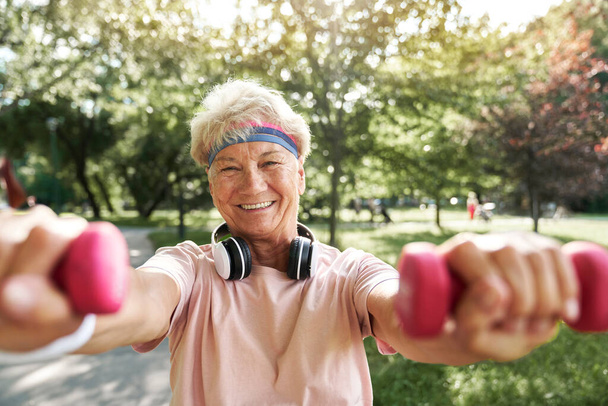 Portrait of senior woman standing in the park and holding weights - Photo, Image