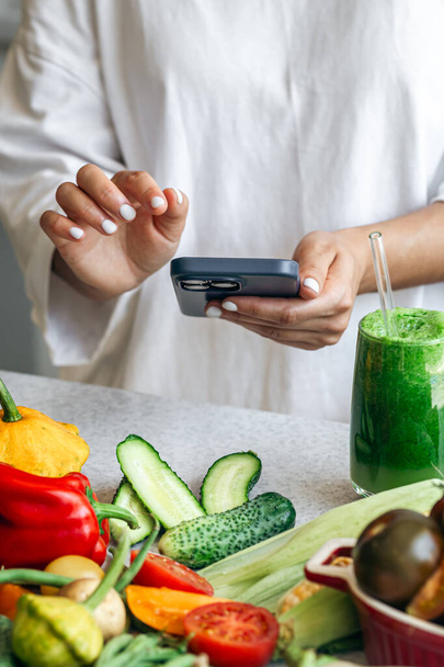 緑色のデトックススムージーを持っているスポーティな女性とキッチンでスマートフォンで栄養アプリを使用して,朝食にフィット. - 写真・画像