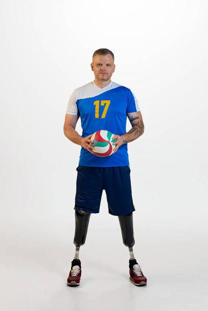 Привлекательный молодой человек с протезной инвалидностью стоя держит волейбольный мяч. Инклюзивный спорт для людей с ограниченными возможностями. Концепция спорта, игрока, медицины, здоровья, ухода за телом. - Фото, изображение