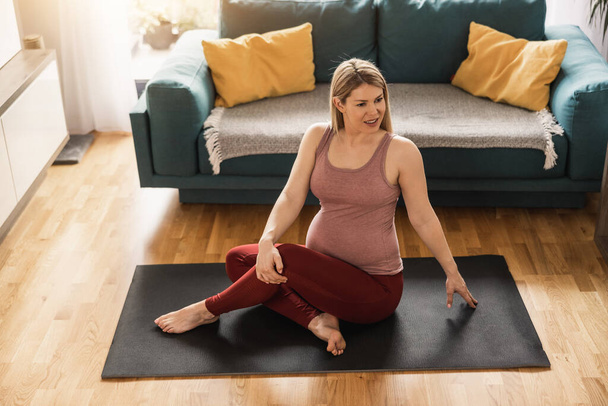 Молода вагітна жінка тренується вдома вранці. Вона тренується на тренувальному килимку і розтягується у своїй вітальні. - Фото, зображення