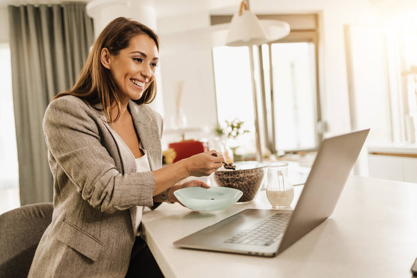 Wielozadaniowa młoda kobieta biznesu korzystająca z laptopa i jedząca śniadanie przygotowując się do pracy. - Zdjęcie, obraz