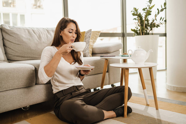 Расслабляющая молодая женщина, сидящая на полу в своей гостиной и наслаждающаяся послеобеденным чаем. - Фото, изображение