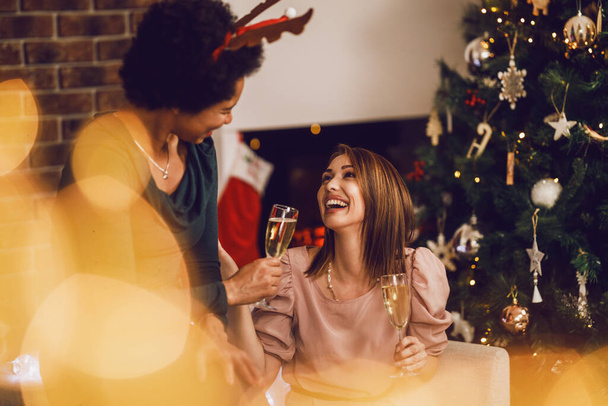 Wesołe wielonarodowe koleżanki bawiące się i wznoszące toast szampanem podczas obchodów Nowego Roku lub Świąt Bożego Narodzenia w domu razem. - Zdjęcie, obraz