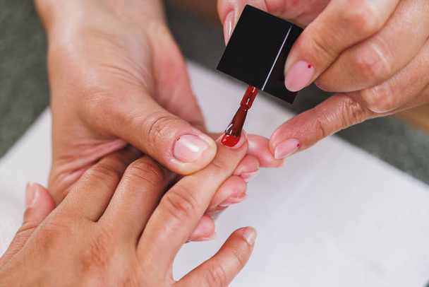 Ausgeschnittenes Bild einer nicht wiederzuerkennenden Kosmetikerin, die während einer Maniküre-Behandlung in einem Schönheitssalon rote Nagellacke auf die Frau aufträgt. - Foto, Bild