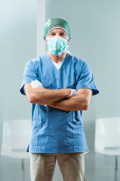 Stolz steht ein robustes Gesundheitspersonal in Krankenhauskleidung, blauem Oberteil, weißen Handschuhen, Mütze und OP-Maske. Ein fitter Mann mittleren Alters, leidenschaftlich für seinen Job, lächelt einen an. - Foto, Bild