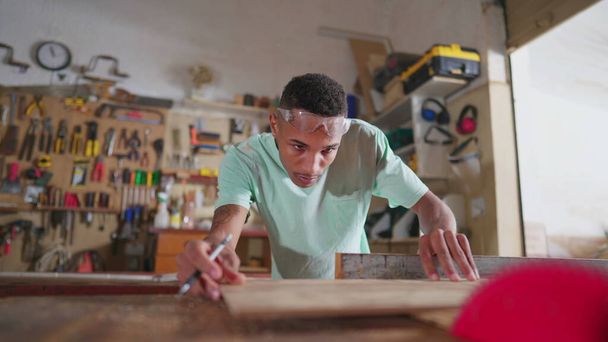 Jeden młody czarny student stolarstwa mierzący drewno, przygotowujący się do użycia pilarki z taśmą pomiarową. Skoncentrowany stolarz w warsztacie, zawód nauczyciela - Zdjęcie, obraz