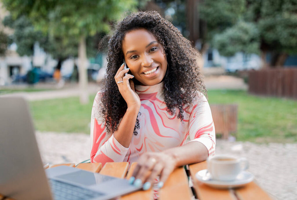 Kaunis nuori musta nainen, joka käyttää kannettavaa tietokonetta ja puhuu matkapuhelimella ulkona kahvilassa, afrikkalainen amerikkalainen freelancer nainen juo kahvia ja ottaa puhelinkeskustelun, istuu pöydässä kahvilassa - Valokuva, kuva