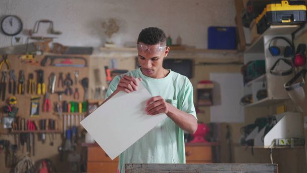Один концентрированный молодой чернокожий бразильский плотник, занятый в мастерской, распиливает древесину с помощью промышленного оборудования. Лицо, занятое работой - Фото, изображение